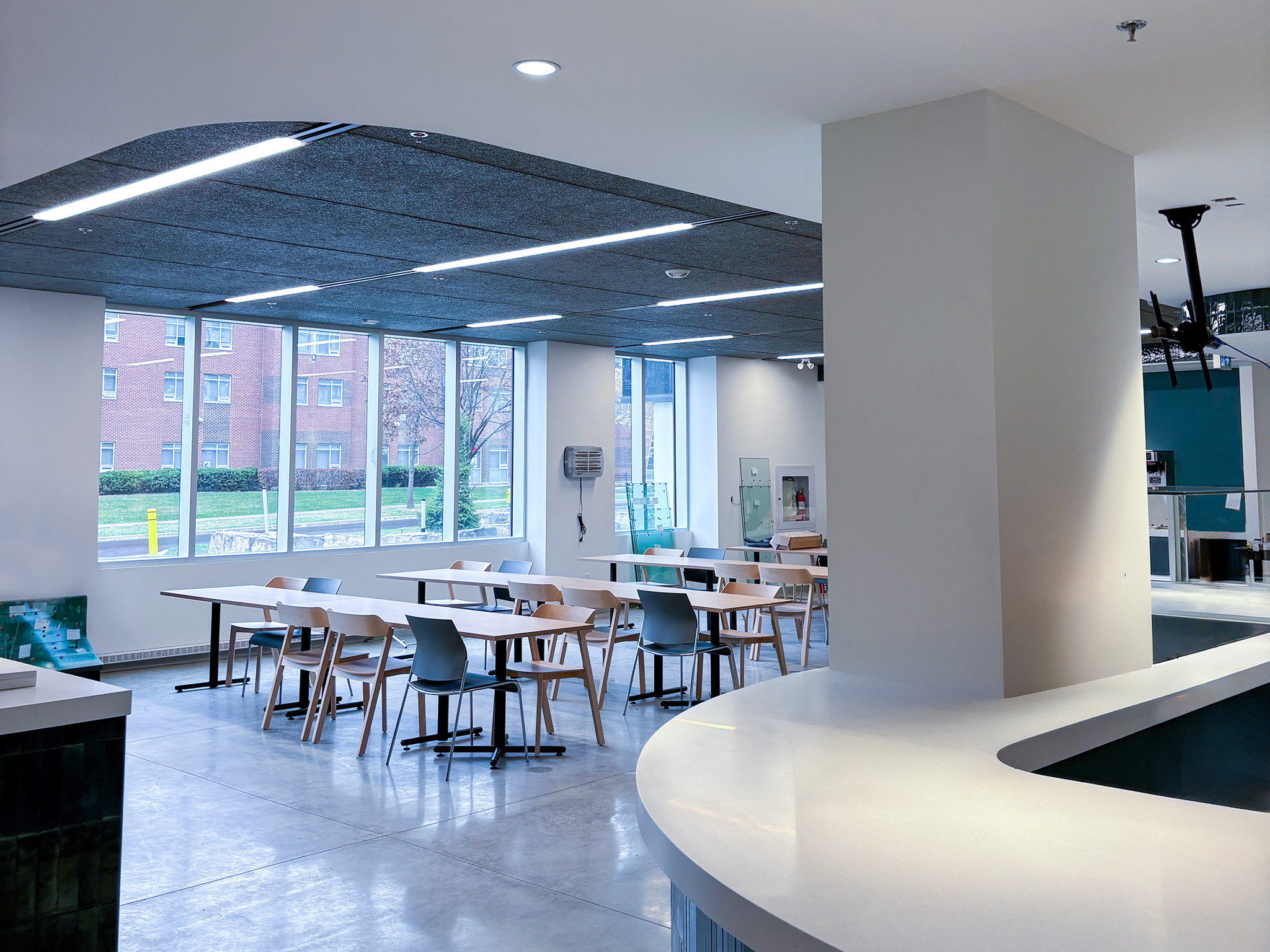 Rodzik Hall (1) Bright dining area with tables, chairs, large windows, recessed lighting, and a curved white service counter.