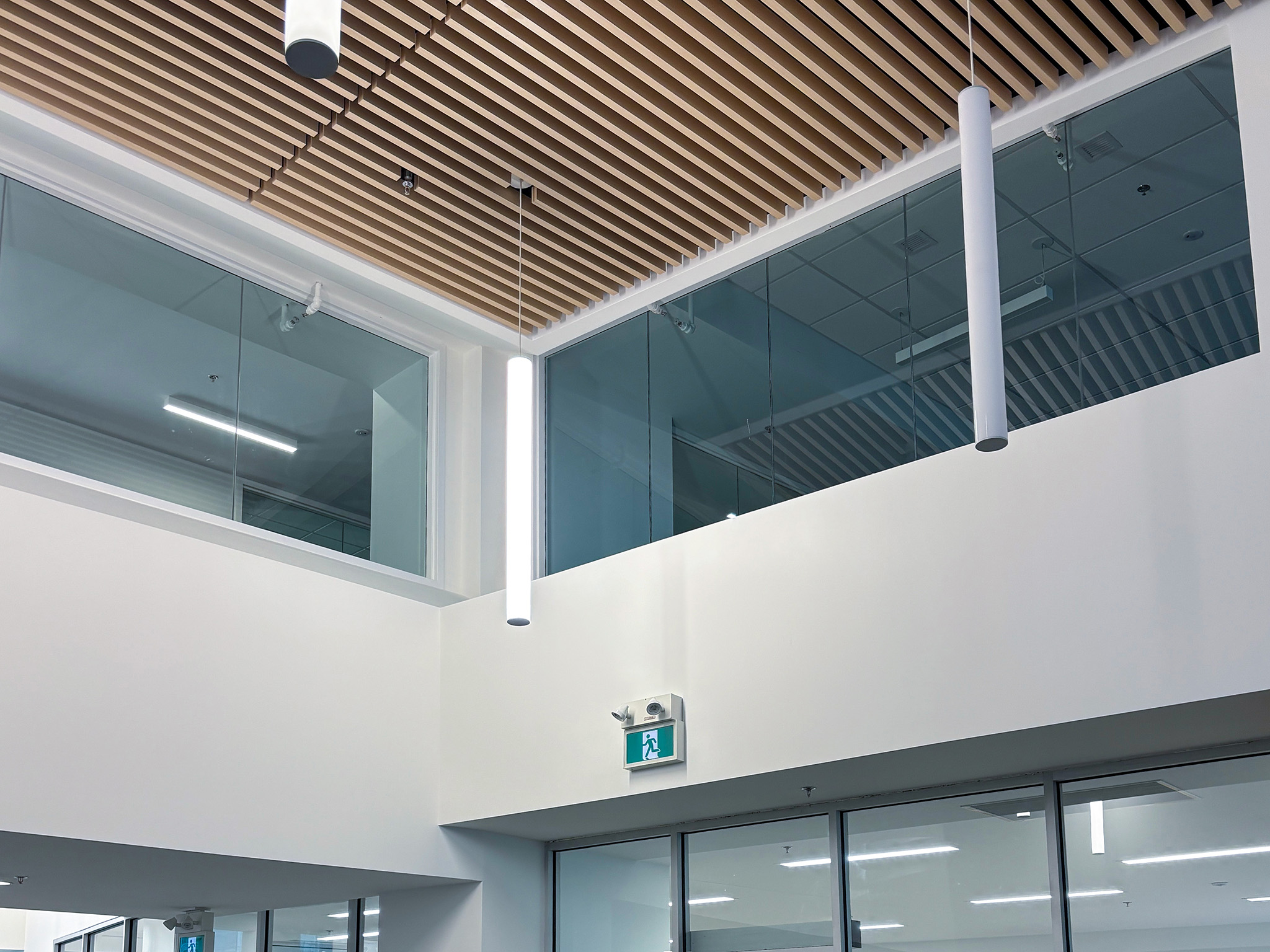 Rodzik Hall (3) Interior view showing a wood-slat ceiling, suspended cylindrical lights, upper-level windows, and an illuminated exit sign.