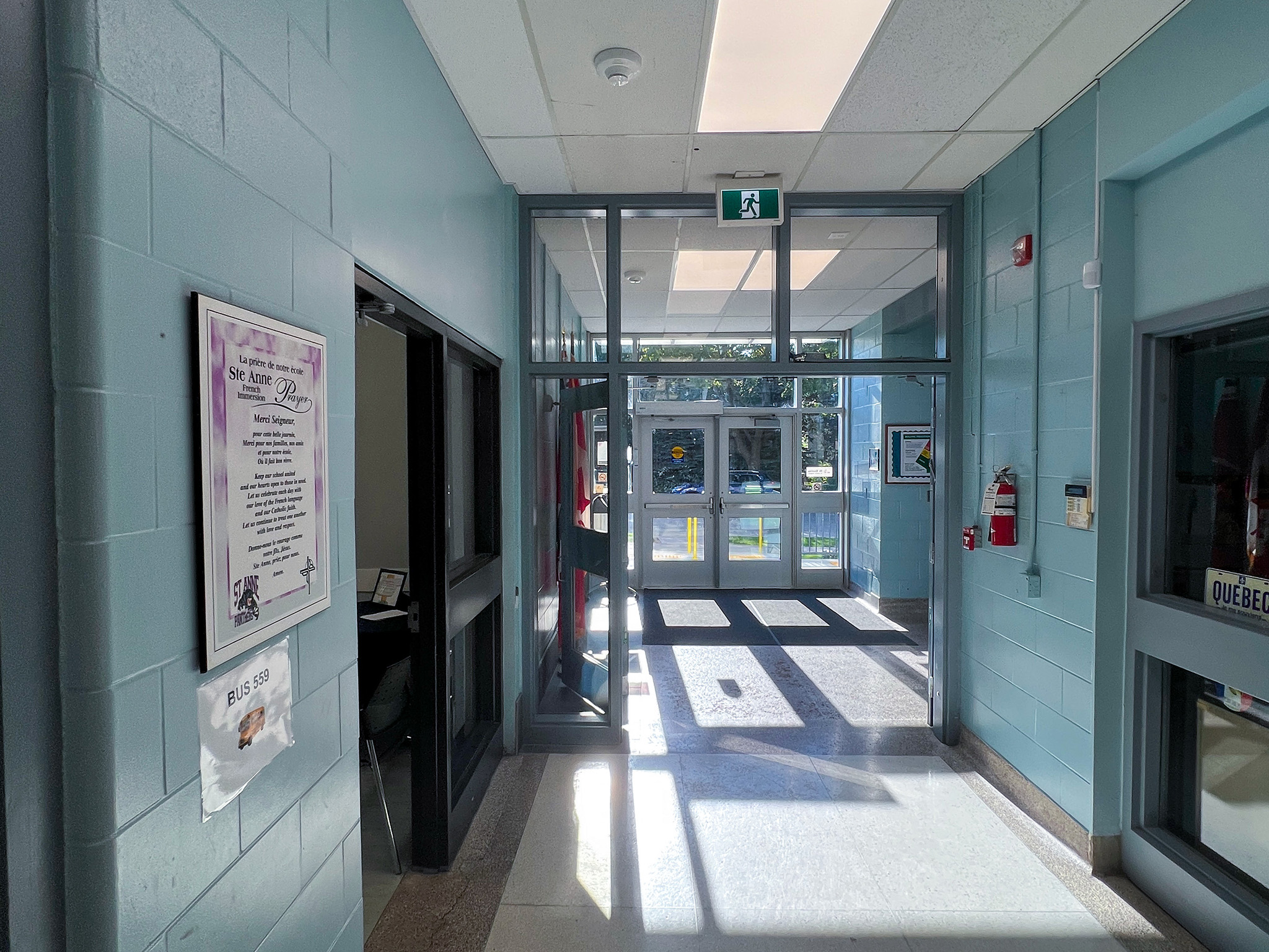 St. Anne French Immmersion Catholic Elementary School (2) School entrance vestibule with glass doors, blue walls, an illuminated exit sign, and sunlight across the floor.