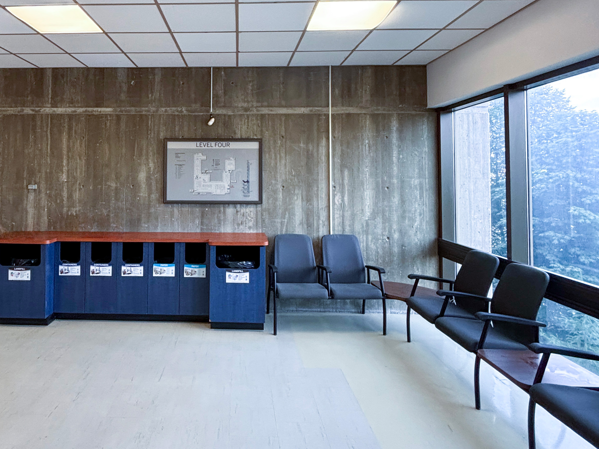 Western University SSC (3) Fourth-floor seating area with blue chairs, recycling bins, exposed concrete walls, and large windows.