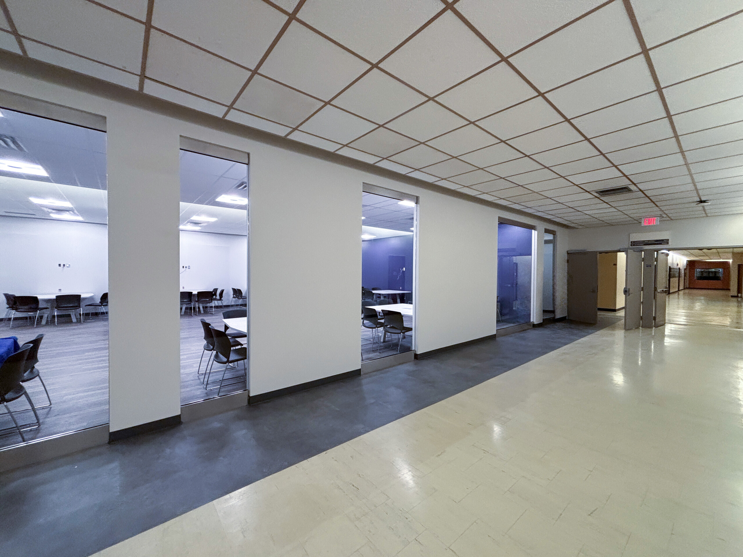 Western University SSC (4) University corridor beside glass-walled learning spaces furnished with tables and chairs, with doors at the far end.
