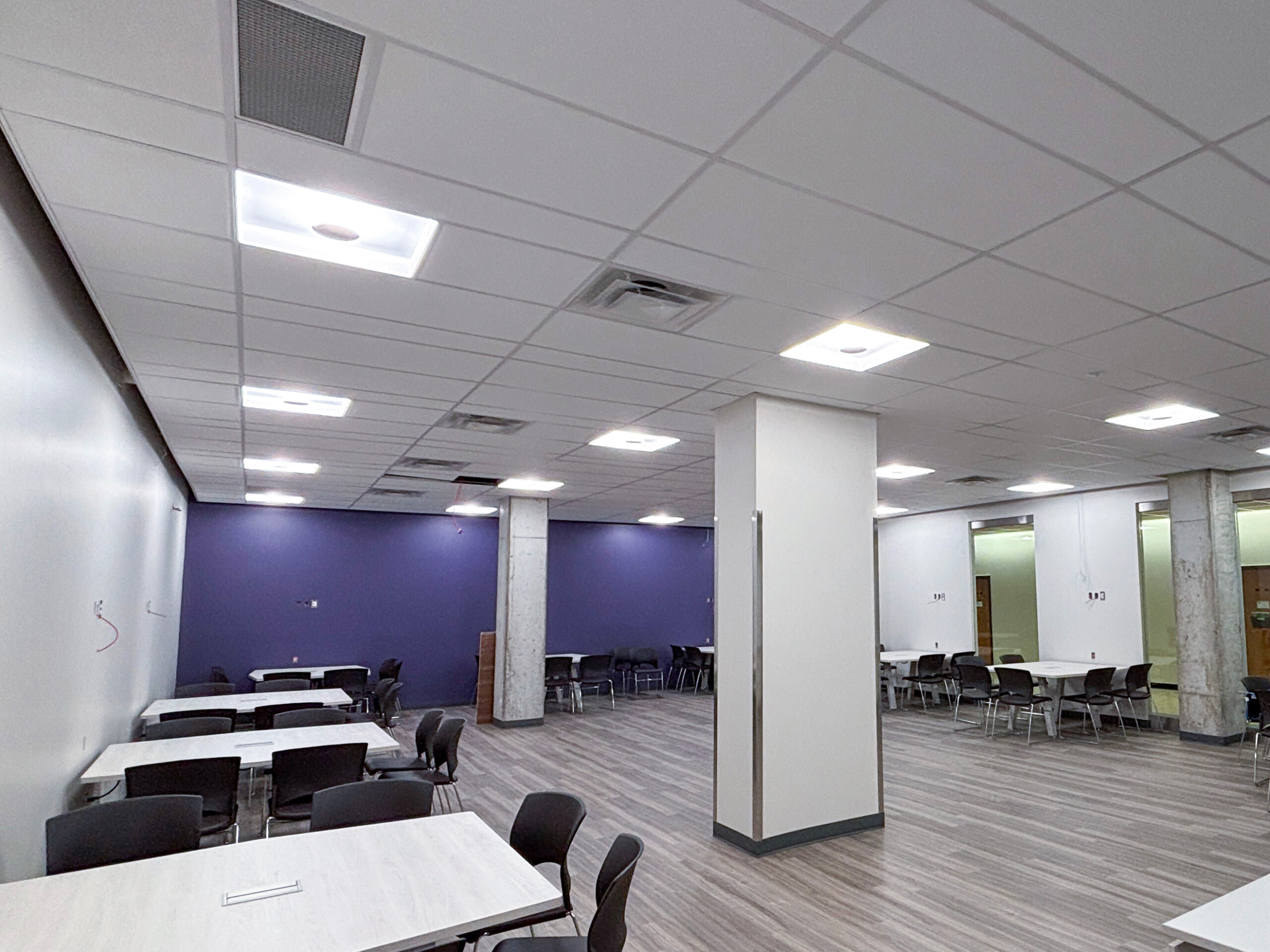 Western University SSC Open university learning space with tables, chairs, purple accent walls, concrete columns, and recessed ceiling lighting.